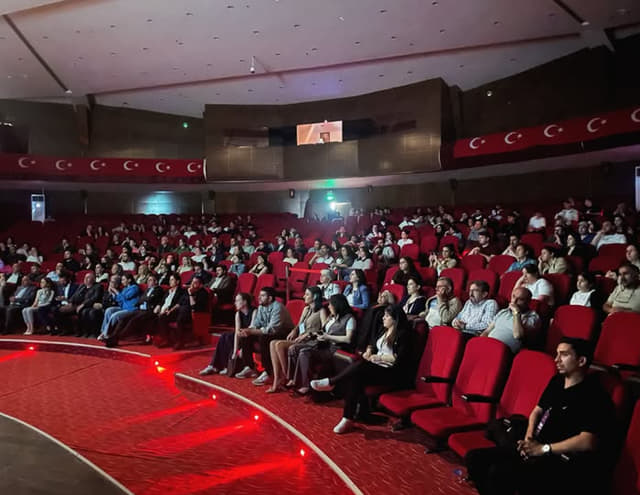 Kültür merkezinin kırmızı koltuklarında oturan kalabalık izleyici topluluğu sahneyi izliyor.